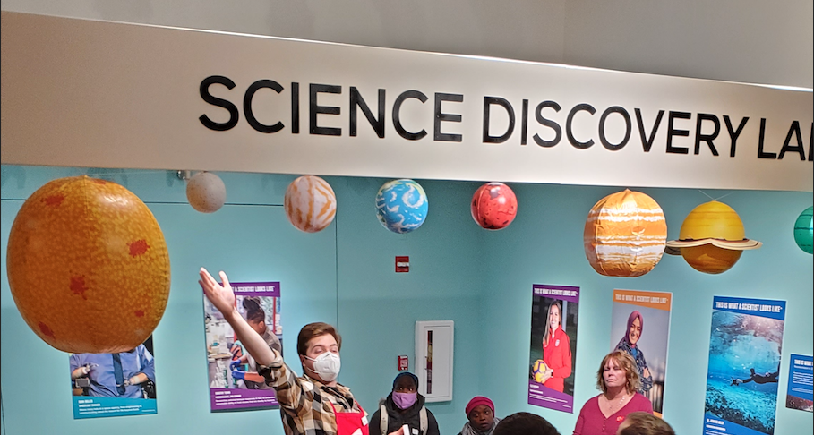 Graham gesticulates wildly in a red apron under a banner reading 'Science Discovery'